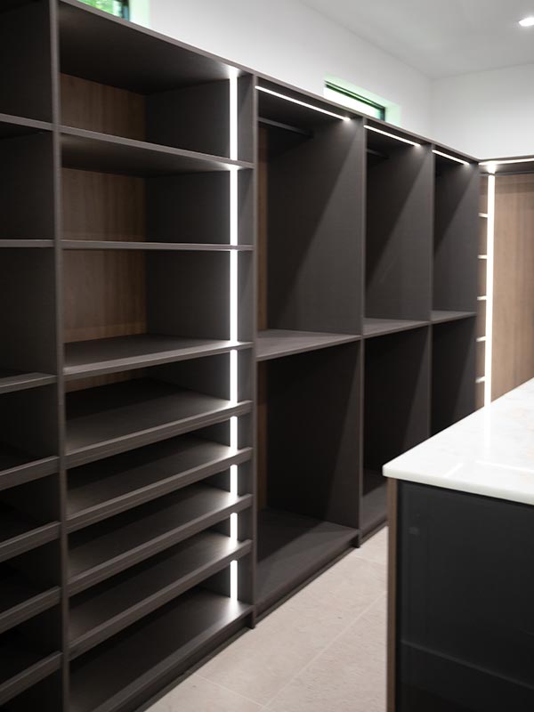 Closet wall of dark graphite shoe shelving with vertical LED strips and open cubbies, with the marble-top island at the right