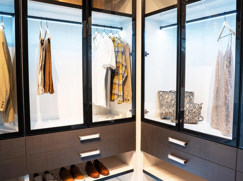 Glass-front closet cabinets with integrated LED lighting, hanging garments, handbags on display and drawers below