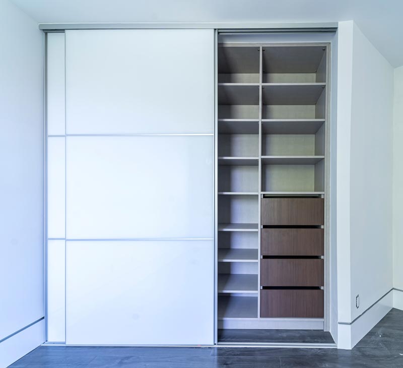Closet de pared con puertas corredizas blancas entreabiertas, con entrepaños ajustables y gavetas de madera oscura