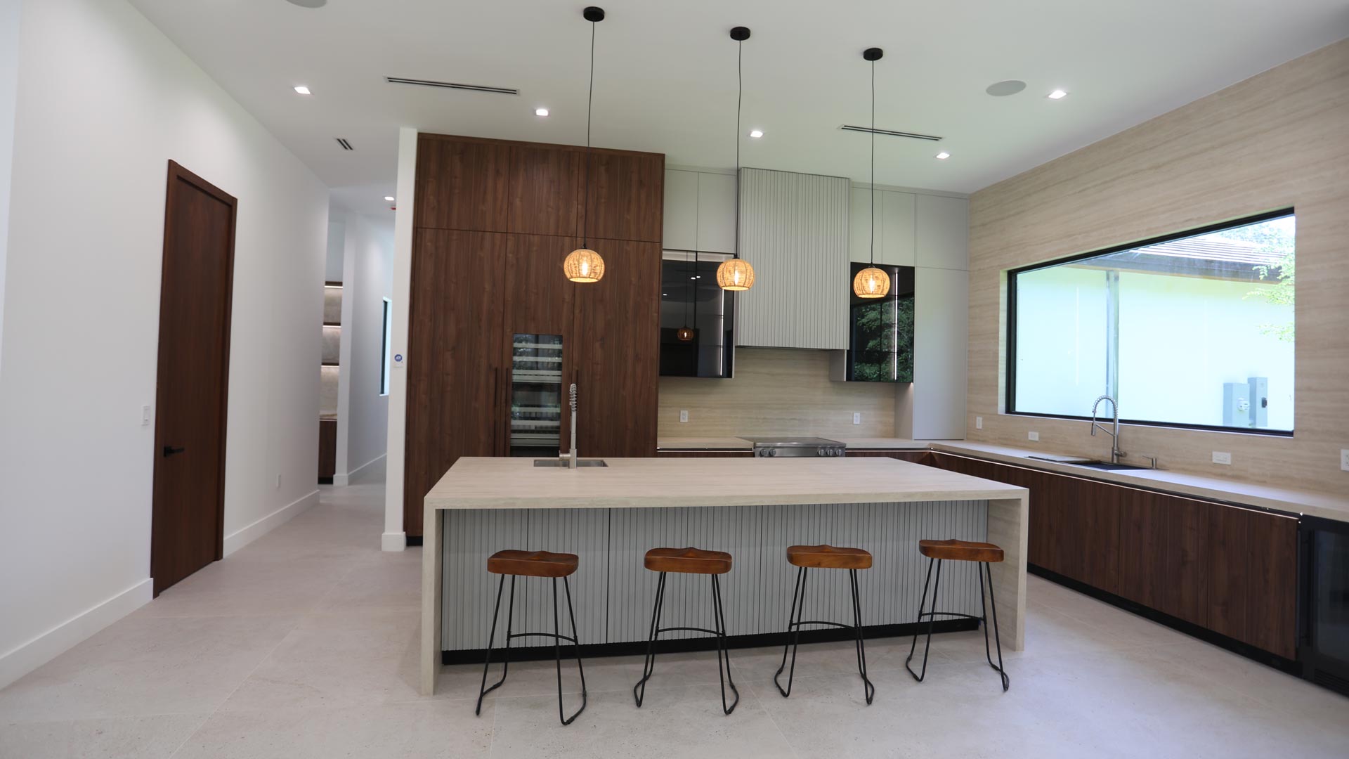 Open-plan custom kitchen in Miami with a travertine waterfall island, bar seating, walnut cabinetry and woven pendant lighting.