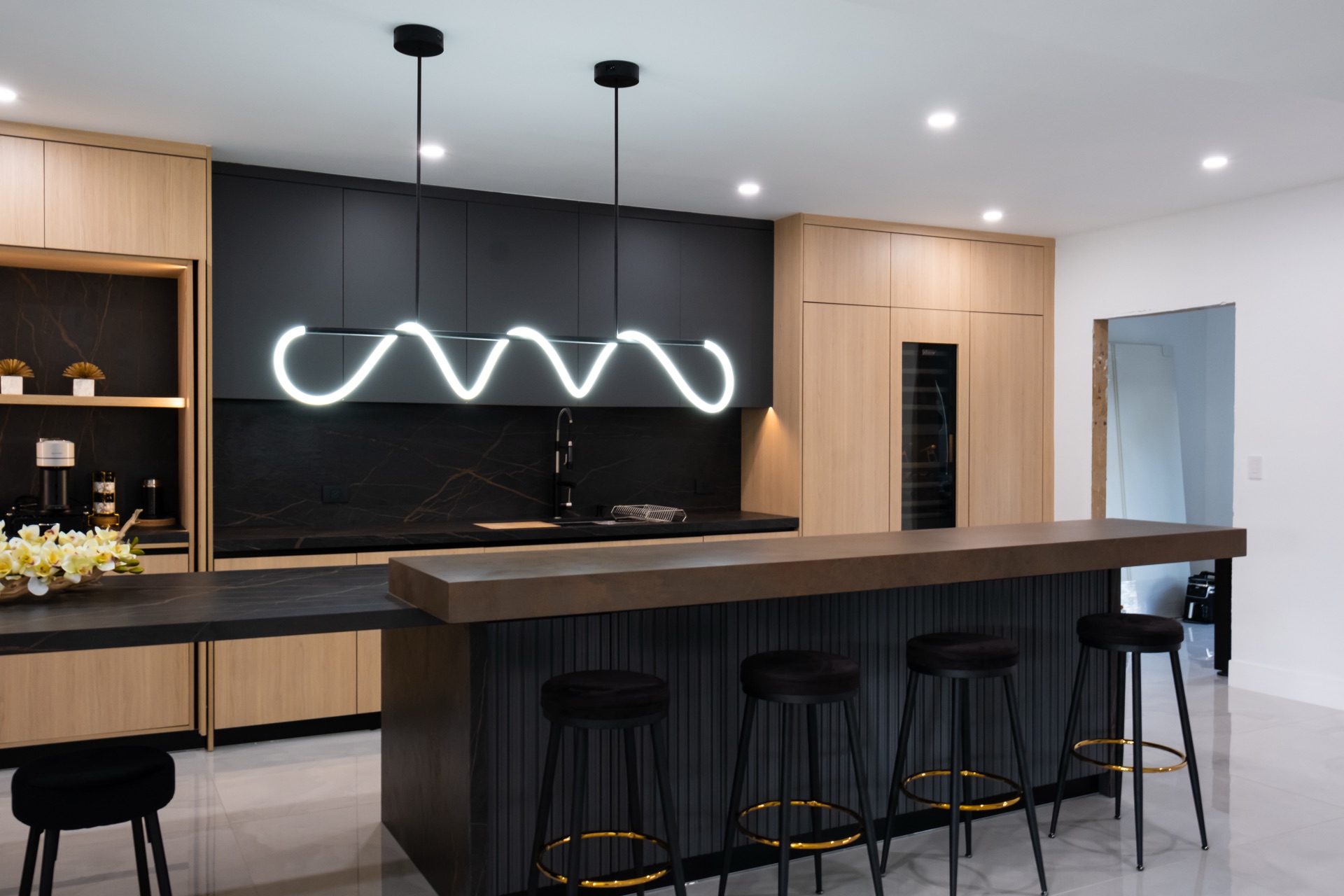 Black and oak kitchen with a bronze-toned island counter, a linear squiggle pendant and four black bar stools along the run.