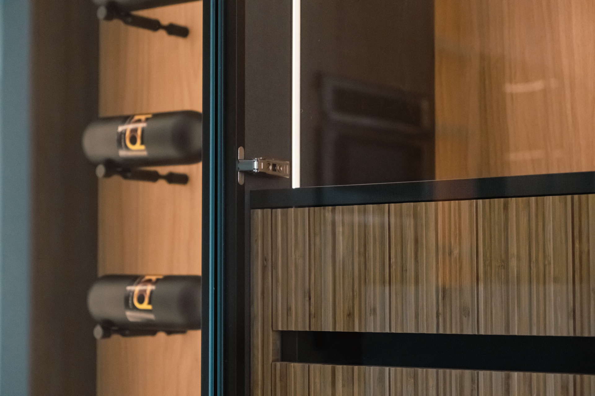 Detail of the concealed hinge on a glass cabinet door, with wine bottles racked behind it and reeded drawer fronts to the right.