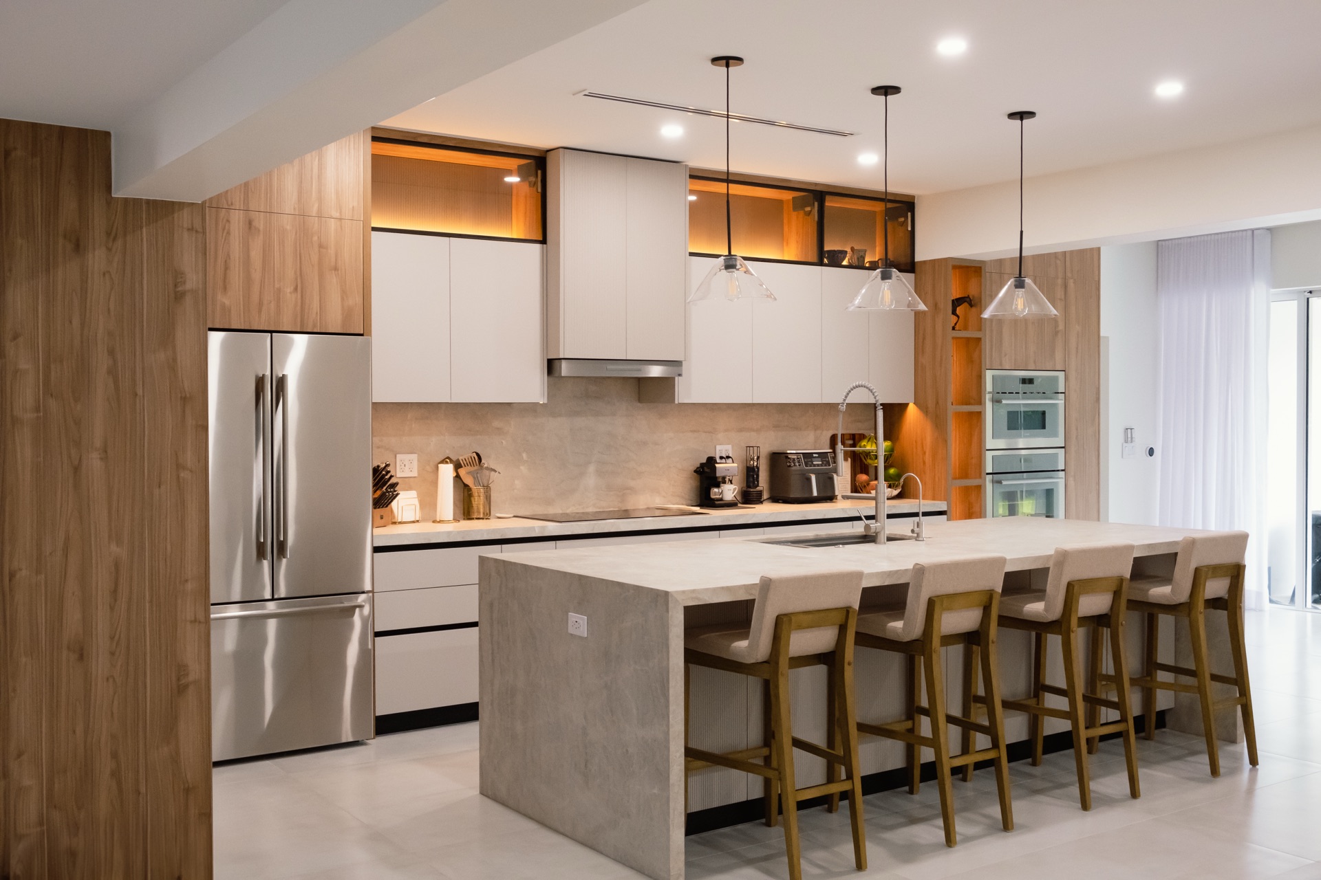 Wide kitchen with a concrete-look island seating four, glass pendants overhead and walnut panelling framing the left side.