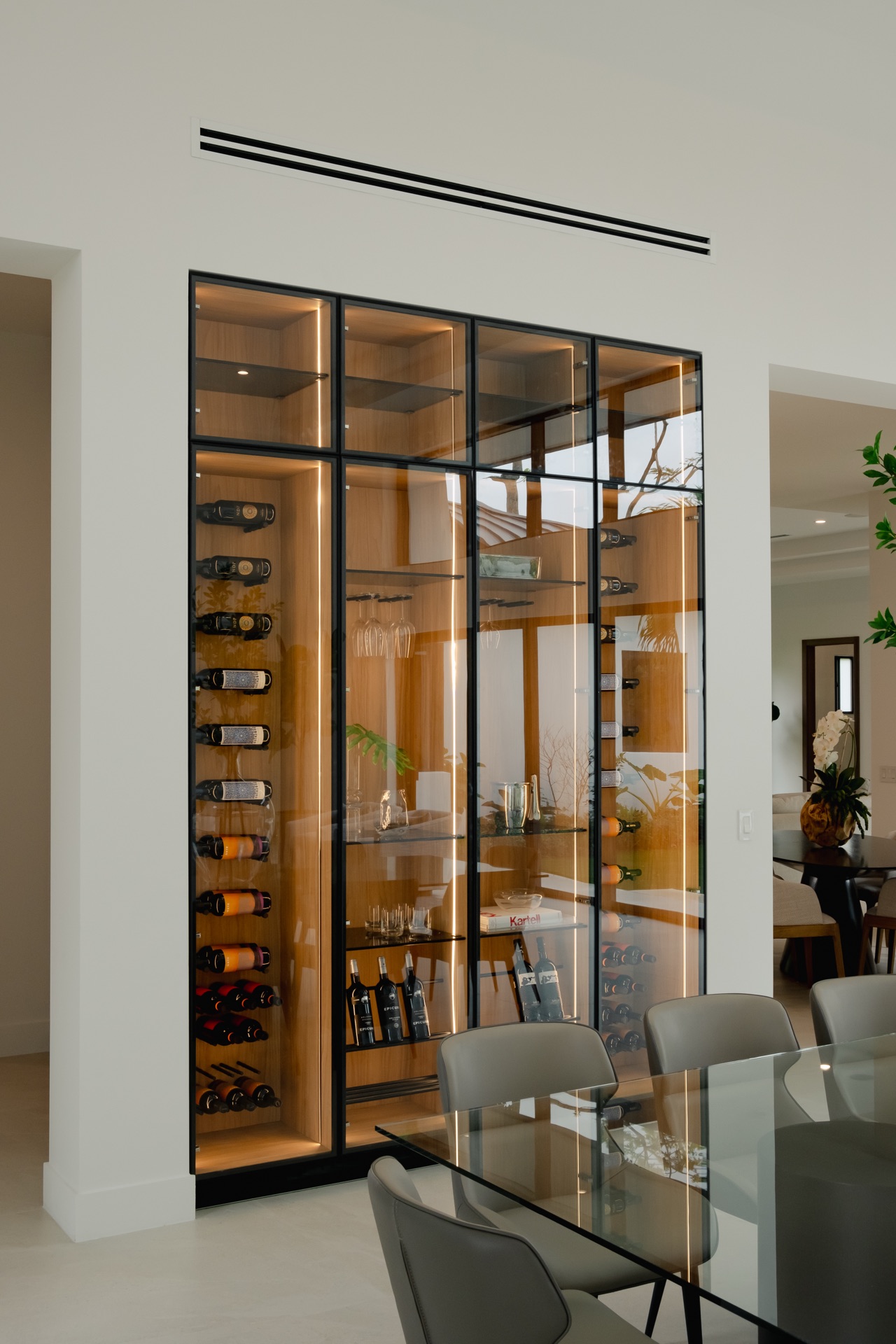 Floor-to-ceiling wine cabinet in black-framed glass, lit from within, standing beside a glass dining table.