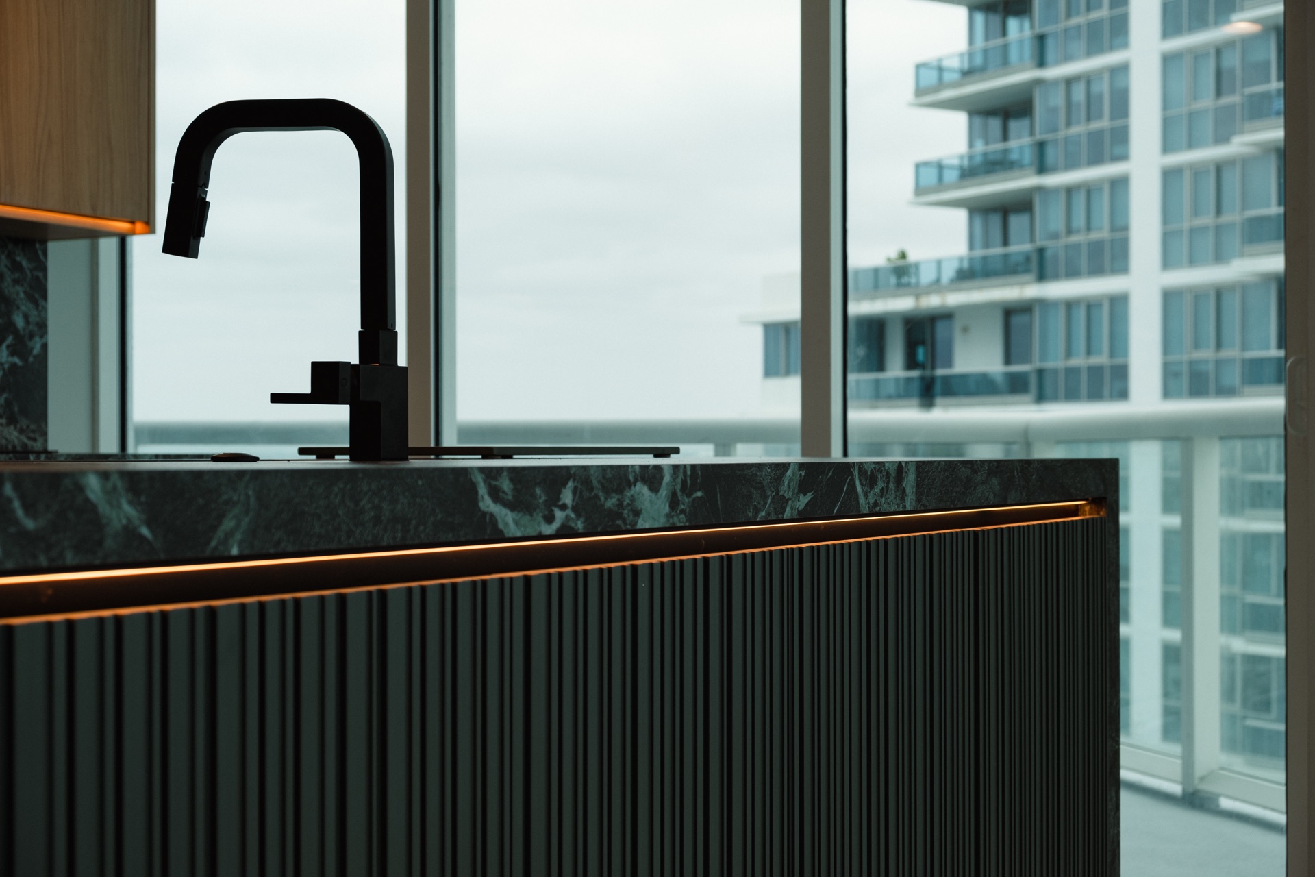 Green marble waterfall island with a black fluted base and a matte black faucet, in front of a window wall overlooking neighbouring towers.