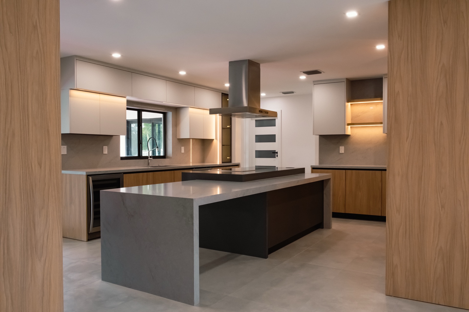 Custom kitchen seen through a walnut-framed opening, with a grey stone island, white handleless upper cabinets and a stainless steel island hood.