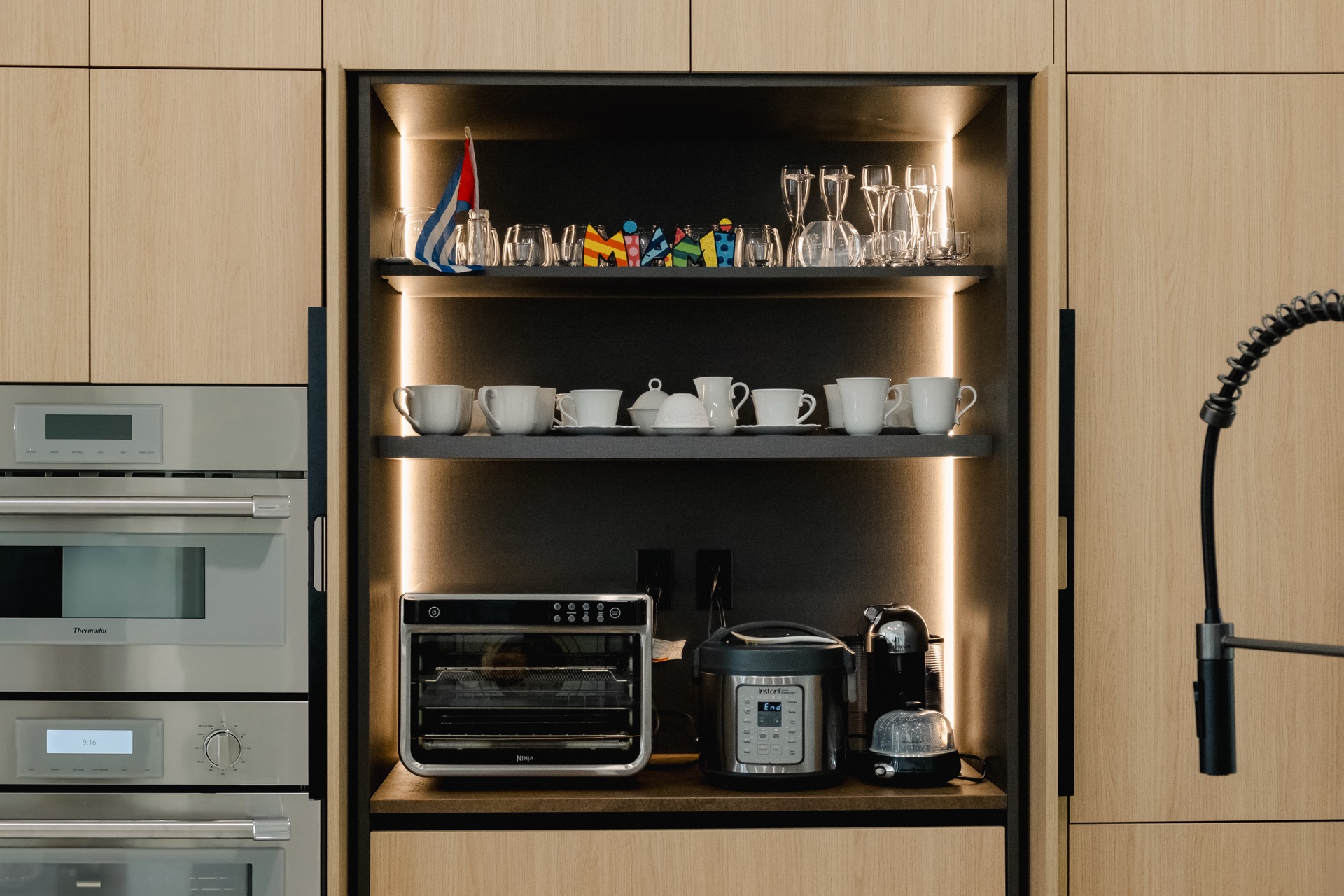 Lit open niche between oak tall units holding cups, glassware and small appliances, next to a stack of built-in ovens.