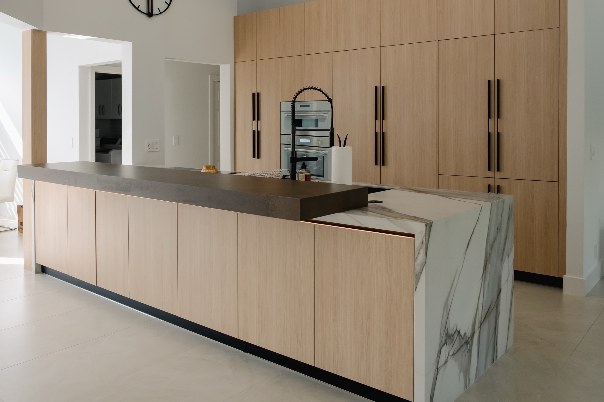 Long oak island topped in brown stone and ending in a white marble waterfall panel, with a full wall of oak tall cabinets and built-in ovens behind.