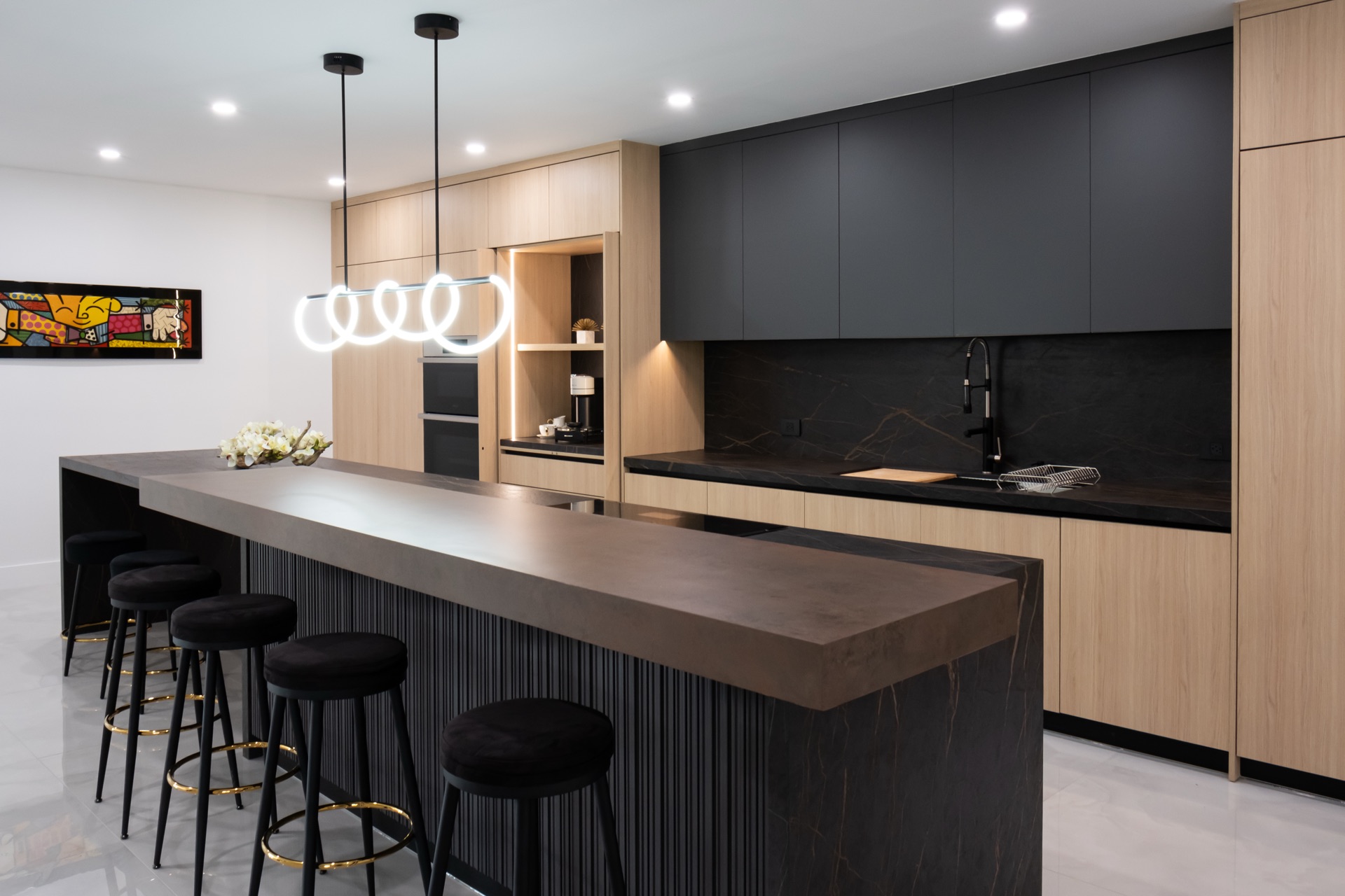 Kitchen in matte black and oak with a bronze-toned island top over a fluted black base, black stools and a ring pendant above.