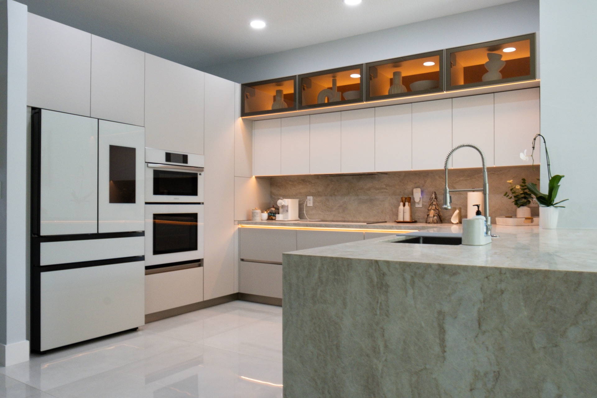 White handleless kitchen with a green quartzite peninsula, stacked built-in ovens and lit glass display boxes above the run.