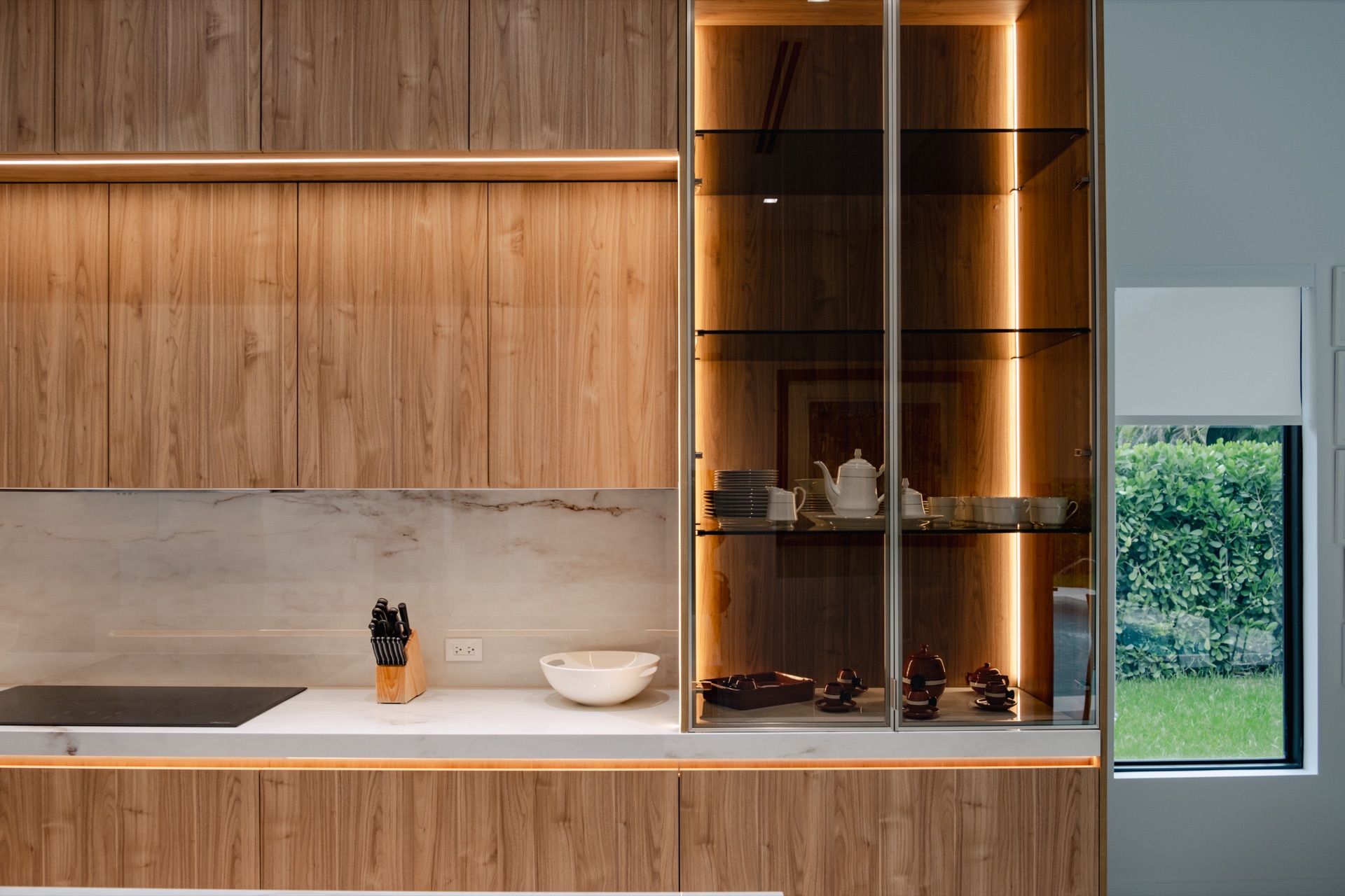Walnut wall panelling above a marble slab backsplash, with a cooktop, a knife block and a lit glass display cabinet on the right.