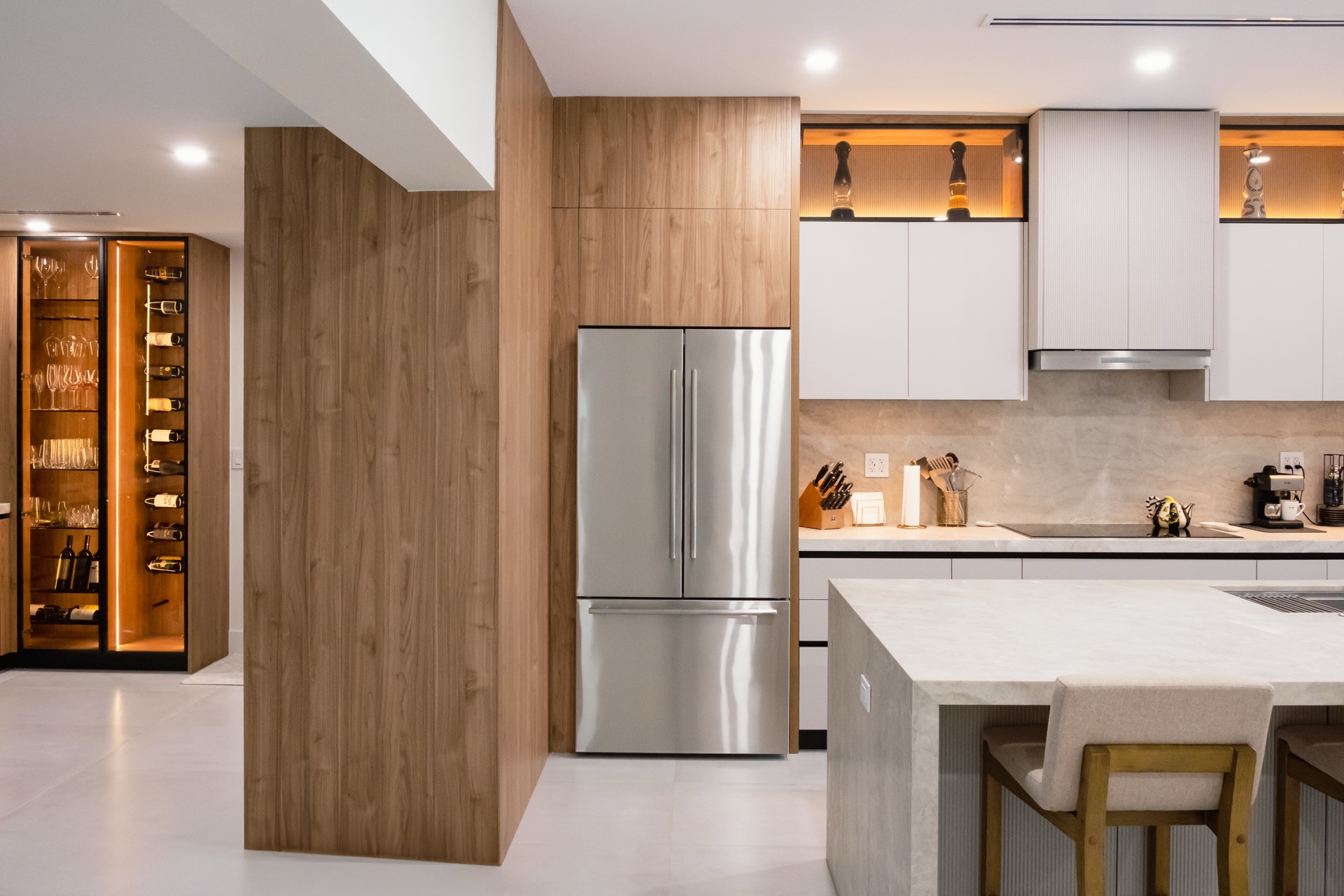 Walnut partition beside an integrated refrigerator column, with a lit wine display to the left and white upper cabinets over the run.