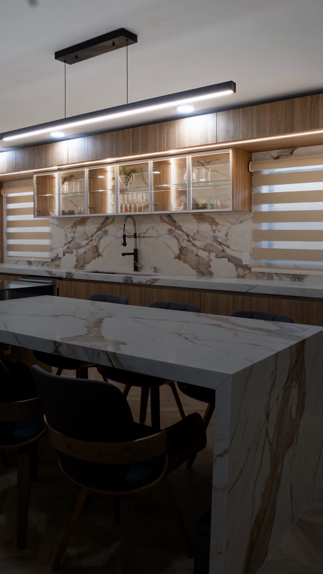 Custom kitchen with LED-lit glass-front display cabinets, walnut cabinetry and a marble waterfall island in warm evening light.